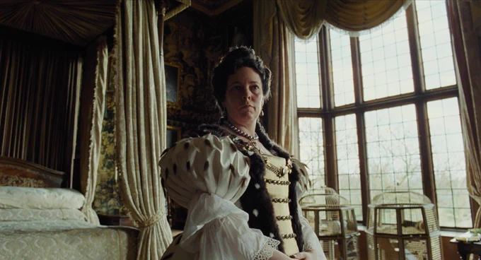 A film still from The Favourite. A woman wears a large dress with big shoulders in a large bedroom. The windows touch the floor and ceilings. She has a serious look on her face as she stares ahead.