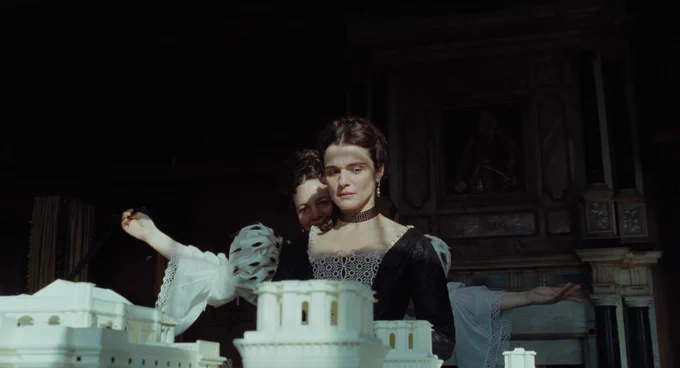 A film still from The Favourite. A woman stands behind another woman, holding her arms out to her sides. She wears a dress with white sleeves and puffy shoulders. The woman in the front wears a darker dress, with her arms to her sides. Her expression is more serious and concerned.