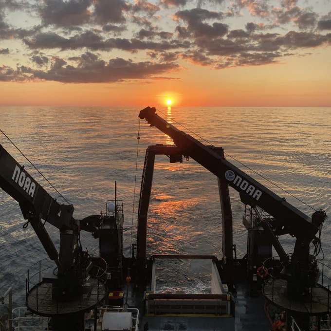 A setting sun shines on the back deck of a research vessel. The vessel has two large cranes on the back deck with the NOAA logo on the sides. The sea is calm, the sky is oranges, golds and blues, and the clouds are gray, peach, pink, and purple.