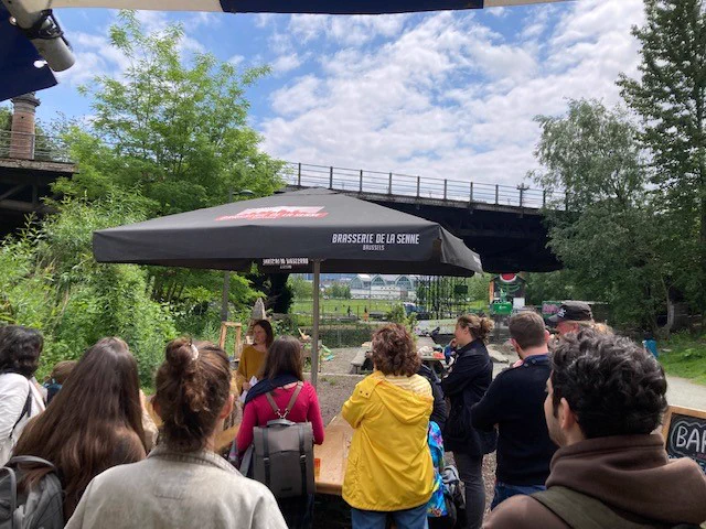 backs of a group of people, outside under an umbrella. There is one person that is facing the camera speaking to the group. In the background there's an industrial bridge, with a city in the background but lots of green space and trees around them.
