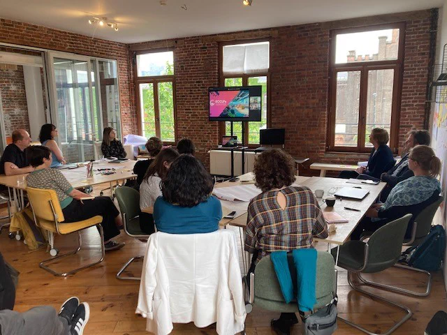 red brick room, people sat around a table, everyone looking at a screen and on it you can see a slide saying ECCLPS and someone speaking