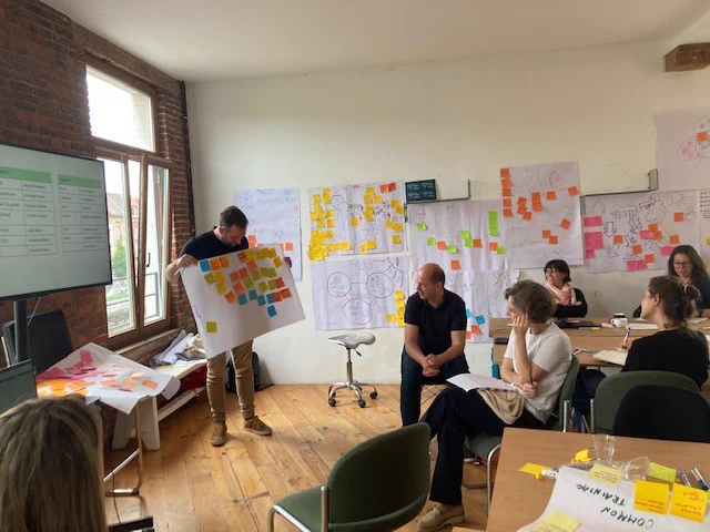 Room with people sat around tables, one person is stood at the front of the room, presenting a big page with lots of post its on it. On the wall behind there's many pages with drawing and post-its stuck on it. 