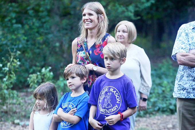 A woman and her three children stand in front of a wooded area.