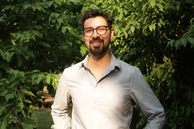 Manuel stands smiling in front of a leafy green tree. He is wearing a white button-down shirt and wearing dark brown glasses.