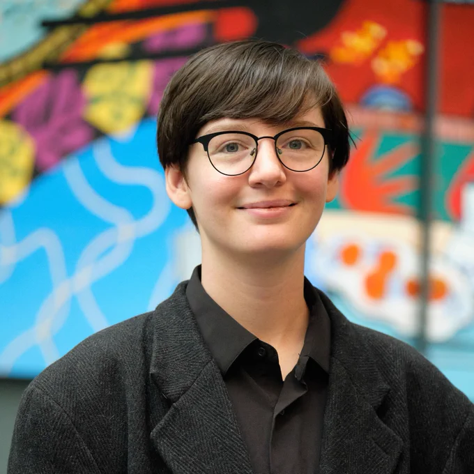 Cel Welch headshot in a coat in front of colorful wall background
