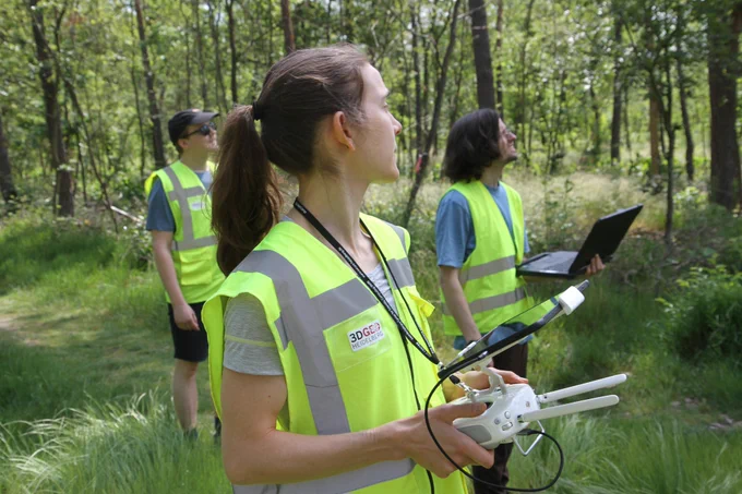 Three people in warn wests looking at the sky towards the same spot. The person in the front has a UAV remote control and tablet in front of her, another person is carrying a field laptop.
