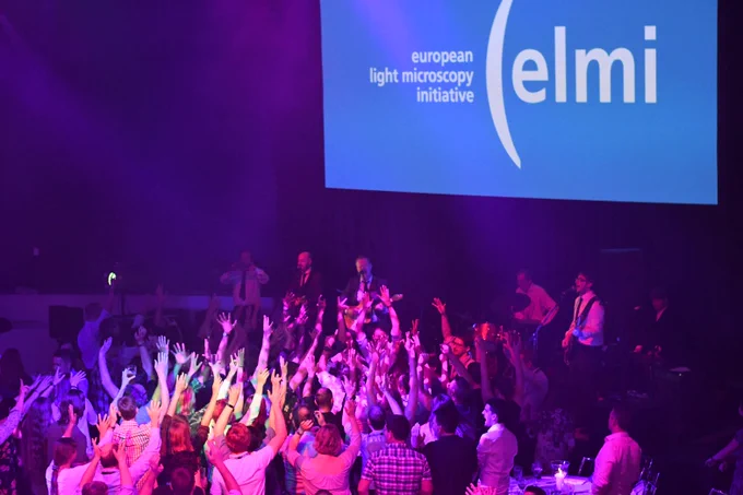 A band plays with a crowded dance floor in the foreground. Large screen behind with elmi logo.