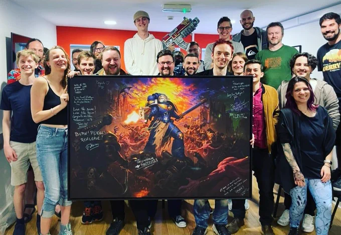 The auroch digital team who worked on Boltgun are stood around holding up a large print of the Boltgun key art signed by members of the Focus team