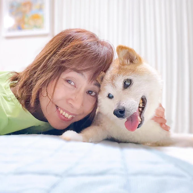 kabosu and her mom pose for a selfie as they press their faces close together and rest on a blanket. kabosu is pretty old here, with some cataracts in her eyes and a mostly white face, but she still looks so happy.