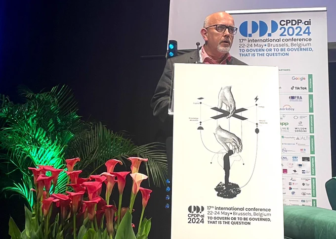 Wojciech Wiewiórowski speaks behind the table and behind him a white banner and on the left side red flowers.