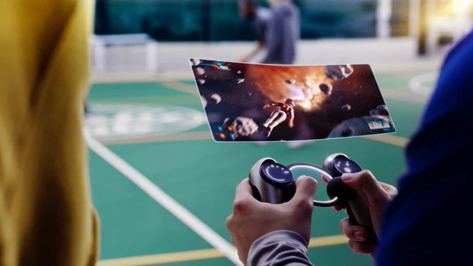 A person’s hands are shown playing a futuristic portable video game console. The controller looks similar to one used for conventional video games, but the screen is floating in midair.