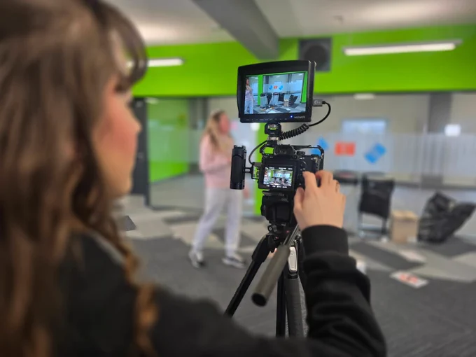 Photo of a person filming another person walk across an empty office. In the background are glass windows and bright green walls. 