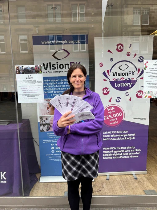 Leanne our operations support manager wearing her purple visionPK fleece. She is stood in front of our shop window with her hand out and a fan of raffle tickets.