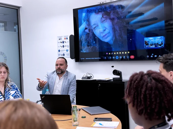 A man talking to the students and a woman can be seen on video call on the tv above 