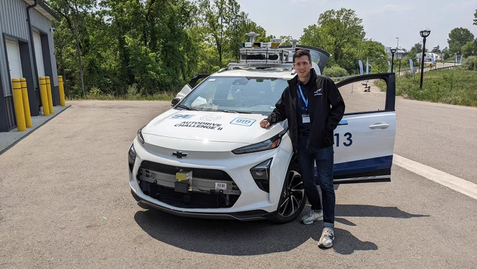 Sandro Papais stands outside, leaning on on the hood of a Chevrolet hatchback that has been converted into an autonomous vehicle. “Autodrive Challenge II” written on the hood of the car and there is a variety of sensors attached to the top of the vehicle.