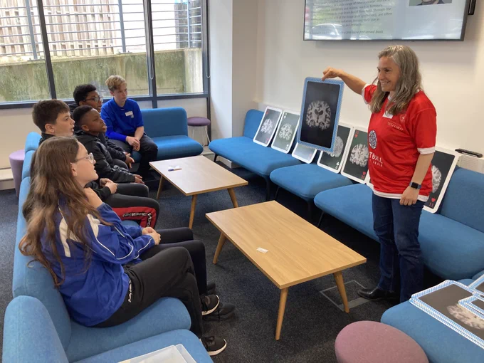Woman in red smiles holding brain scan image up to group of smiling teens in black and blue on blue couches