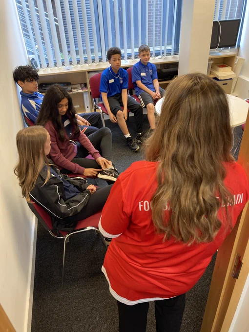 Brunette woman in red shirt stands in doorway facing away from camera towards teen students sitting in chairs