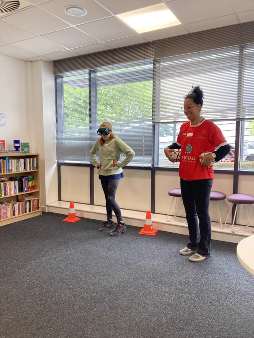 Woman in red stands holding mini footballs while girl wearing prism goggles stands hands on hips next to her between orange cones