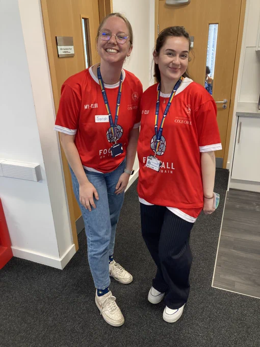 Two women in red shirts and Oxford Uni staff lanyards smile at the camera