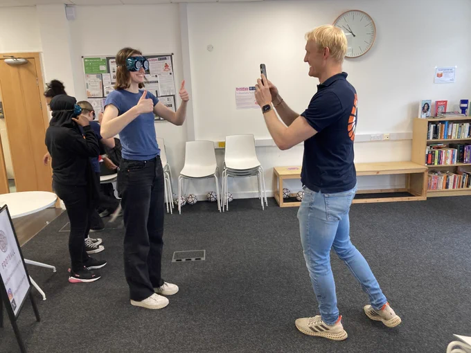Woman in blue wears prism goggles and gives thumbs up to man in blue taking her photo