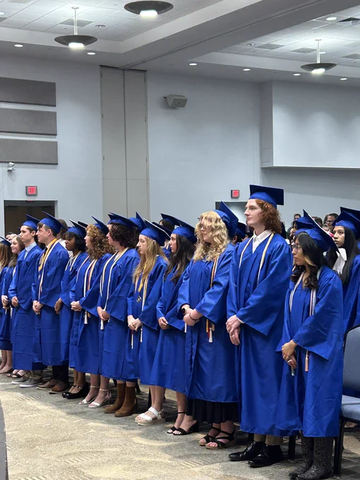 About 20 graduates in the front row standing at their chair.