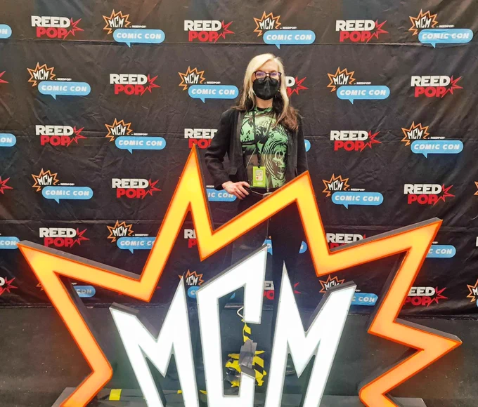 Zoë standing behind the glowing MCM sign.
She has long hair and is wearing a black and green top and black blazer.