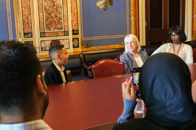 Senator Murray is sitting at a large red table, looking to the left and smiling at a boy in suit who is one seat away from her. To the right of the Senator, a woman in a white cardigan is sitting. Across the table, two people are sitting with their backs to the camera, the person on the right is holding a phone with the camera app open.