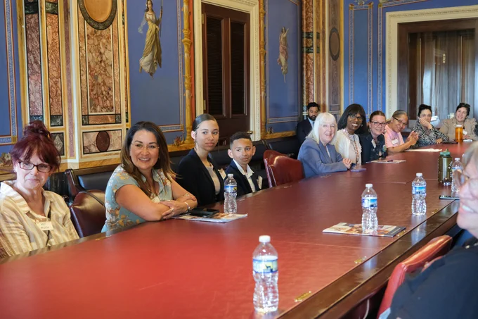Senator Murray is sitting at a large red table with 10 other people pictured. On the table there are multiple water bottles and pamphlets. Everyone pictured is looking to the left of the frame.