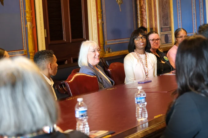 Senator Murray is sitting at a red table, smiling and looking to the right of the frame. There are three women sitting to the right of her, and one boy sitting to the left. There are tow women sitting across from them with the back of their heads facing the camera.
