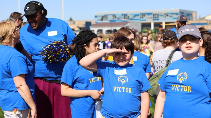 A group of individuals wearing t-shirts with different inscriptions, 'VICTOR' and 'VOLUNTEER,' at an outdoor event. One child, in the center, wearing a 'VICTOR' shirt and a cap, shields their eyes from the sun while looking towards the distance. The adults and other children around appear engaged and joyful, conversing and smiling under clear skies.