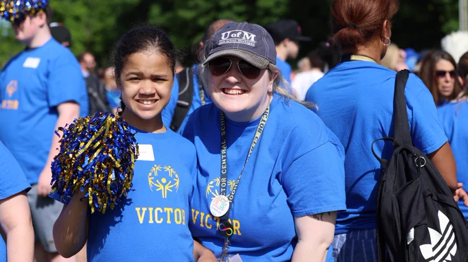 Two people in blue t-shirts, one wearing a medal and holding a pom-pom, smiling at an outdoor event with other attendees in the background. Their shirts have "VICTOR" printed on them.