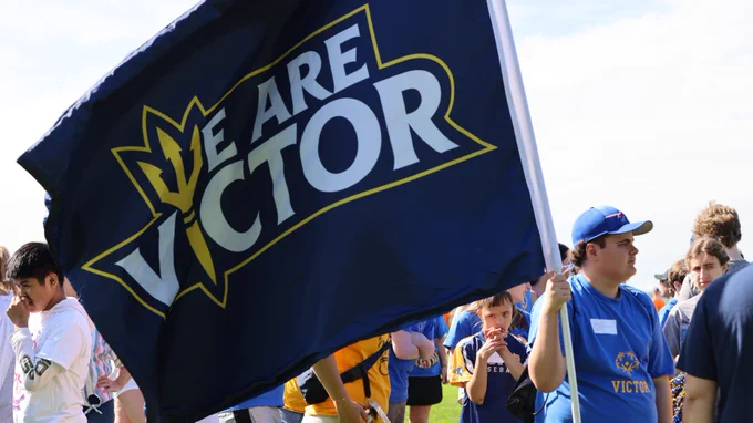 A group of individuals participates in an outdoor event, prominently displaying a large "We Are Victor" banner under a clear, sunny sky.