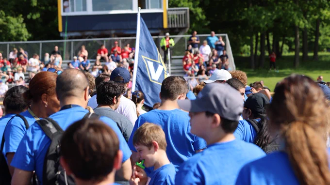A group of people in blue t-shirts at a community event outdoors, with a flag displaying the Villanova University logo at the forefront, surrounded by spectators in front of a small building.