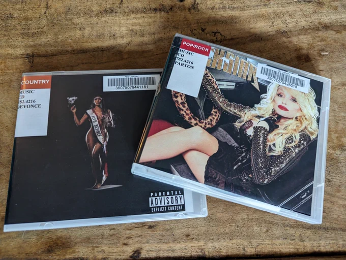 Library copies of Beyonce's Cowboy Carter and Dolly Parton's Rockstar on CD just vibing together on a wood table.