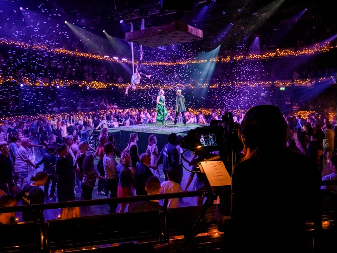 Two people stand on a stage as a person is lowered down towards them. A crowd of people watch on. A person stands next to a camera, recording.