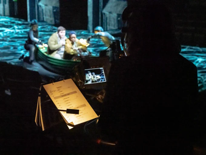 A boy and a girl sit in a boat. They are filmed through a camera pointing towards a stage.