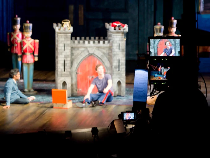 A man sits in front of a small toy castle. A camera points towards him.