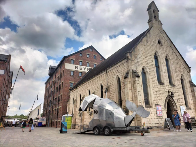Big metal whale on wheels next to an old church. In the distance are sailing ships.