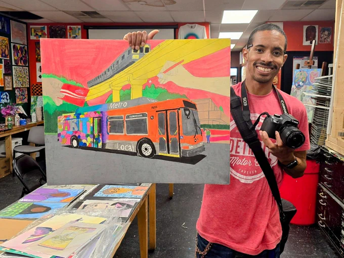 A man wearing a pink shirt holds a colorful artwork of a bus. He stands in an art classroom, smiling, with a camera around his neck.