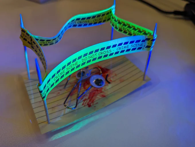 A tiny crime scene diorama. The victim is Clippy the Microsoft Office assistant, lying dead in a pool of blood. Wait, do paperclips have blood?