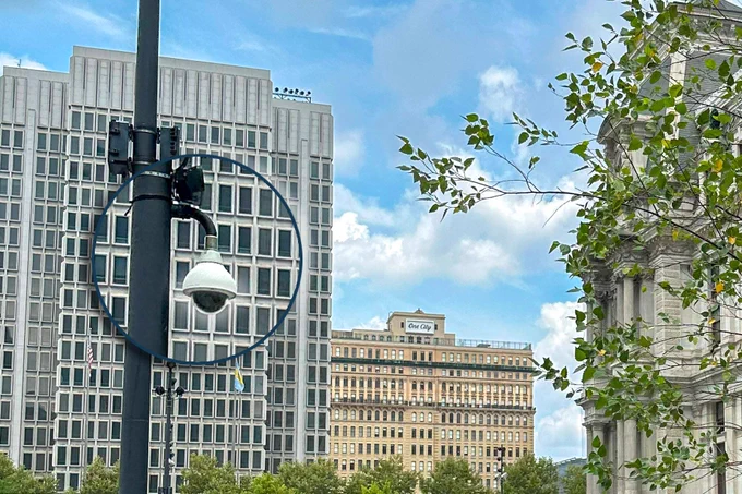 A security camera mounted on a pole in front of modern office buildings and a tree with green leaves, under a partly cloudy sky.
