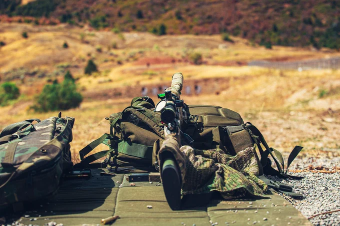 A sniper rifle with a mounted scope, camouflaged with fabric and netting, lies atop a green mat on a gravel ground. Nearby, a backpack and other tactical gear are arranged on the ground. This setup is positioned in a natural landscape with a dry, hilly background.