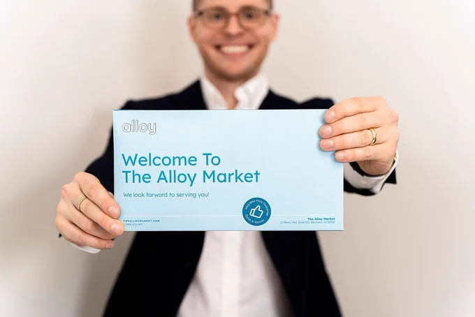 Person in a suit holding a sign that reads, "Welcome to the Alloy Market. We look forward to serving you!" The person is smiling and has their hands slightly blurred in the foreground.