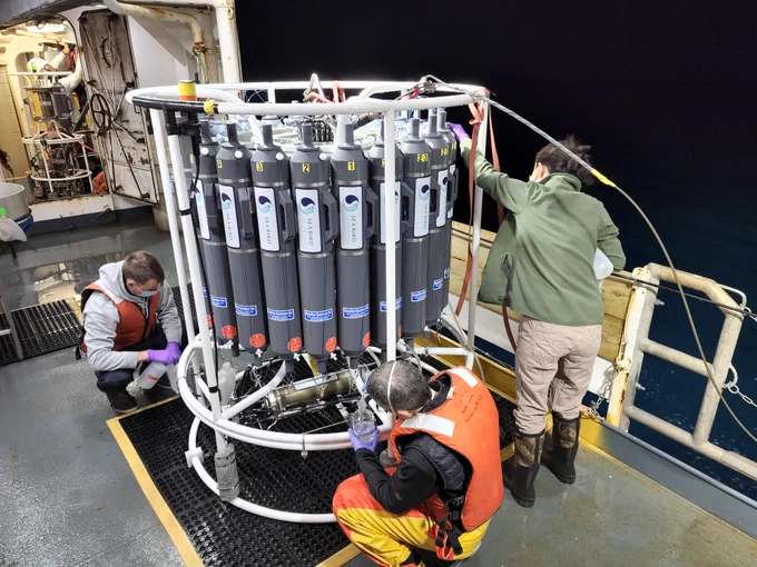 Three scientists collect water samples from a large piece of ocean sampling equipment. The equipment sits on the side deck of a research vessel and consists of a white metal circular frame that is about six feet tall. The frame has several vertically-placed gray bottles attached to it that the scientists are collecting water from via tubing.