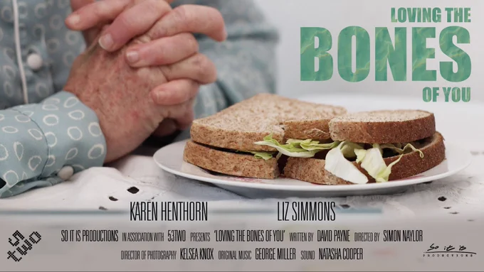 An image of a lady’s clasped hands. She wears a blue/green blouse with white circles on. We can just see her hands in front of a sandwich in brown bread, on a white plate, on a white table cloth. The title reads ‘Loving The Bones Of You’.