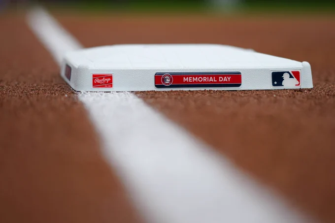 Third base rests planted in the infield of a baseball diamond. It has Rawlings and Major League Baseball logos and a Memorial Day 2023 Base Jewel in center. The left field foul line, blurred, runs vertically through the photo.
