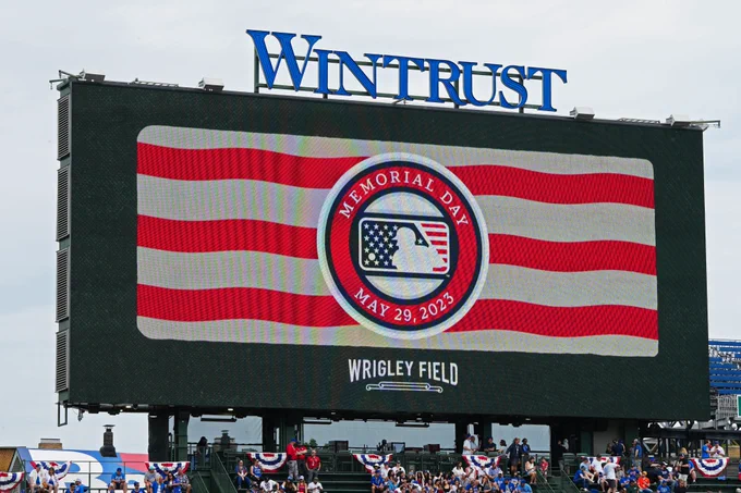 Wrigley Field's left field Video Board displays a Memorial Day 2023 graphic, featuring red and white stripes and the MLB logo mocked up as an American flag with stars and stripes around it.