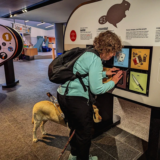 Museum exhibition, a woman at a panel, touching an interactive display, a guide dog behind her.