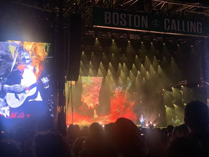 Ed Sheeran performing at night at Boston Calling. He’s small and barely recognizable. Ed is front and center with a guitar. Colorful oranges, red and yellow lighting / imagery are on a large screen in the back. To the front there is a massive screen with Ed on it and it’s intentionally styled to be washed out but still cool looking.