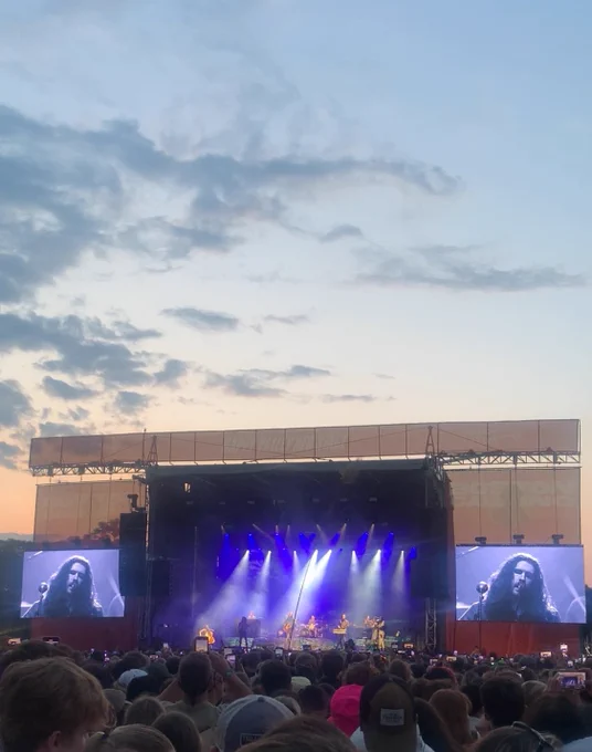 Hozier playing at Boston Calling as the sunset sets over the festival stage. There’s yellow / orange sunset colors with evening clouds rolling in. Hozier is in the middle of the stage. Barely recognizable at this distance. Blue/purple and white overhead lights shine from the top over him and his band. Two large screens flank the stage where you can see Hozier’s face and his distinctive long hair. He’s crooning into the song “Would That I” chorus that has an awesome line, “it’s not tonight… that I hold you tight…”